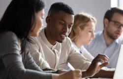African and asian different age colleagues seated at desk in co-working room look at laptop screen discuss new corporate app, more experienced mate explain program software to intern mentoring concept